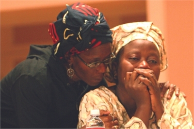 [ai] Two women seated at a table, one gently leaning on the other, who has her hands clasped in front of her face. They are indoors, with a soft-focus background, conveying a moment of support or contemplation.